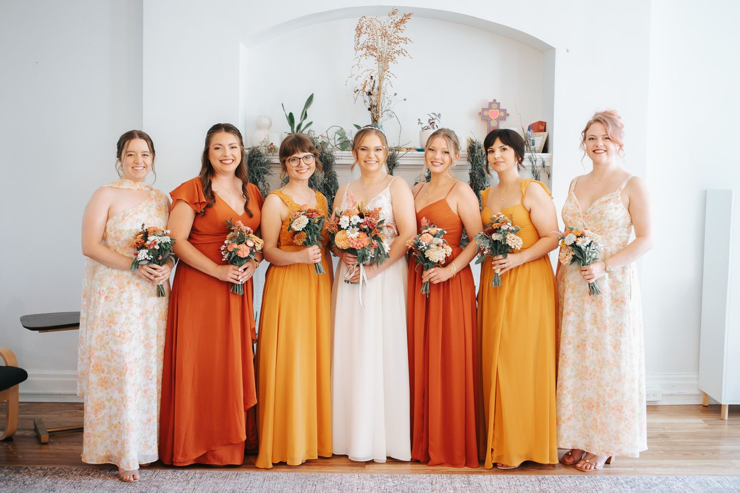 Seven bridesmaids in rust, orange, and floral dresses hold small bouquets while posing together in a bright room with an arched alcove.
