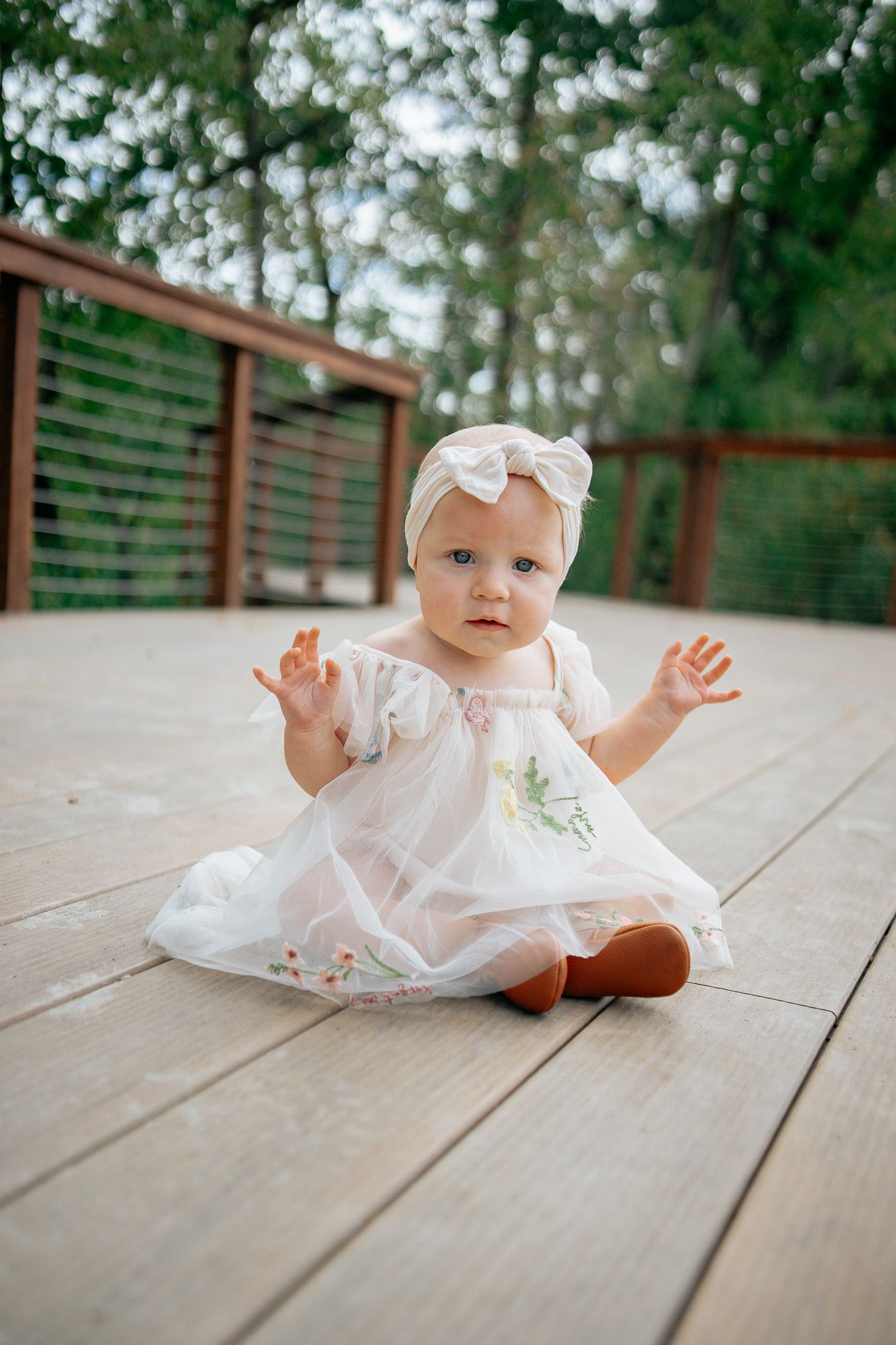 A baby in a white dress lays on a wooden deck while reaching up with both hands.