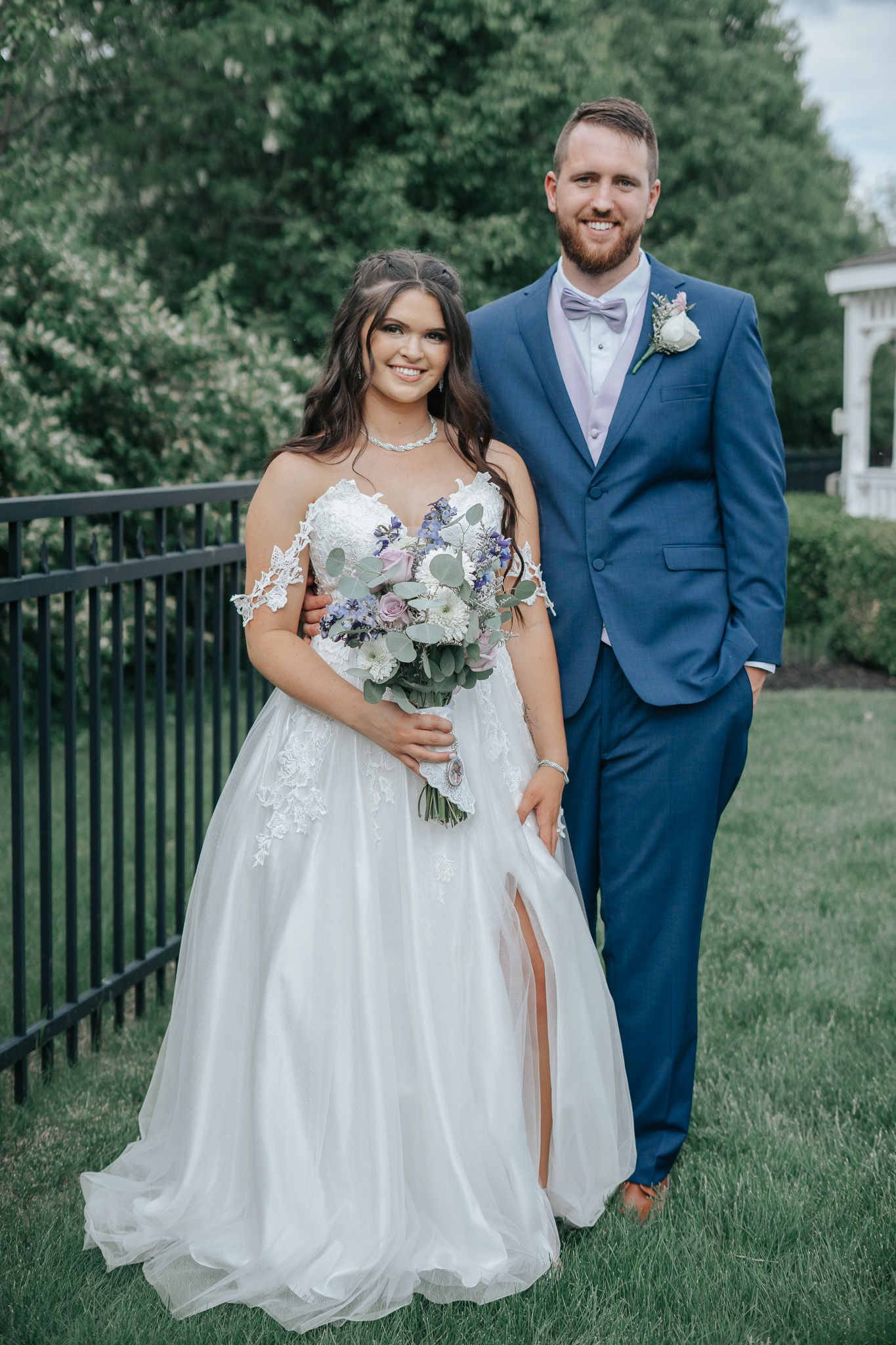Bride in white lace gown holding floral bouquet stands with groom in blue suit on green lawn.