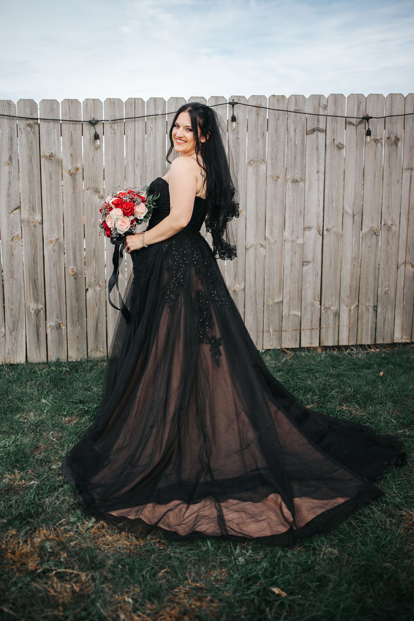A person in a dramatic black lace wedding gown poses with a red bouquet against a wooden fence.