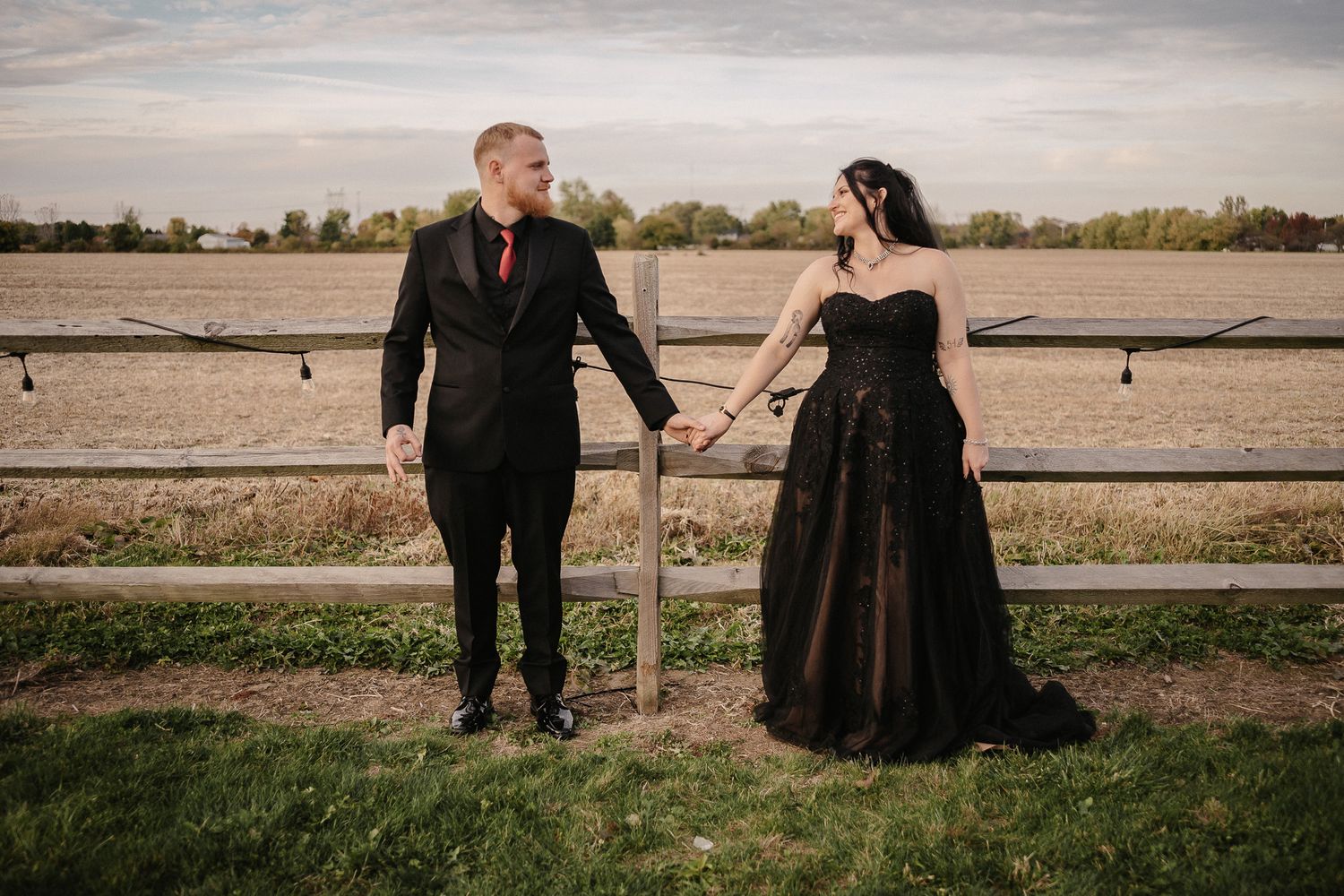 Couple in matching black wedding attire holding hands by a rustic fence in an open field.