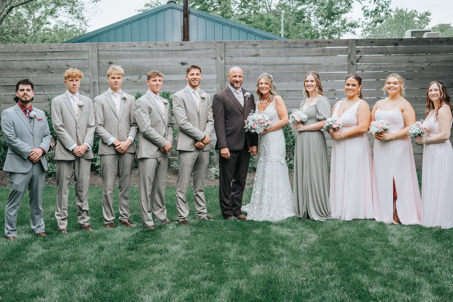 Wedding party in gray suits and sage green bridesmaids dresses pose together on lawn in front of wooden fence.