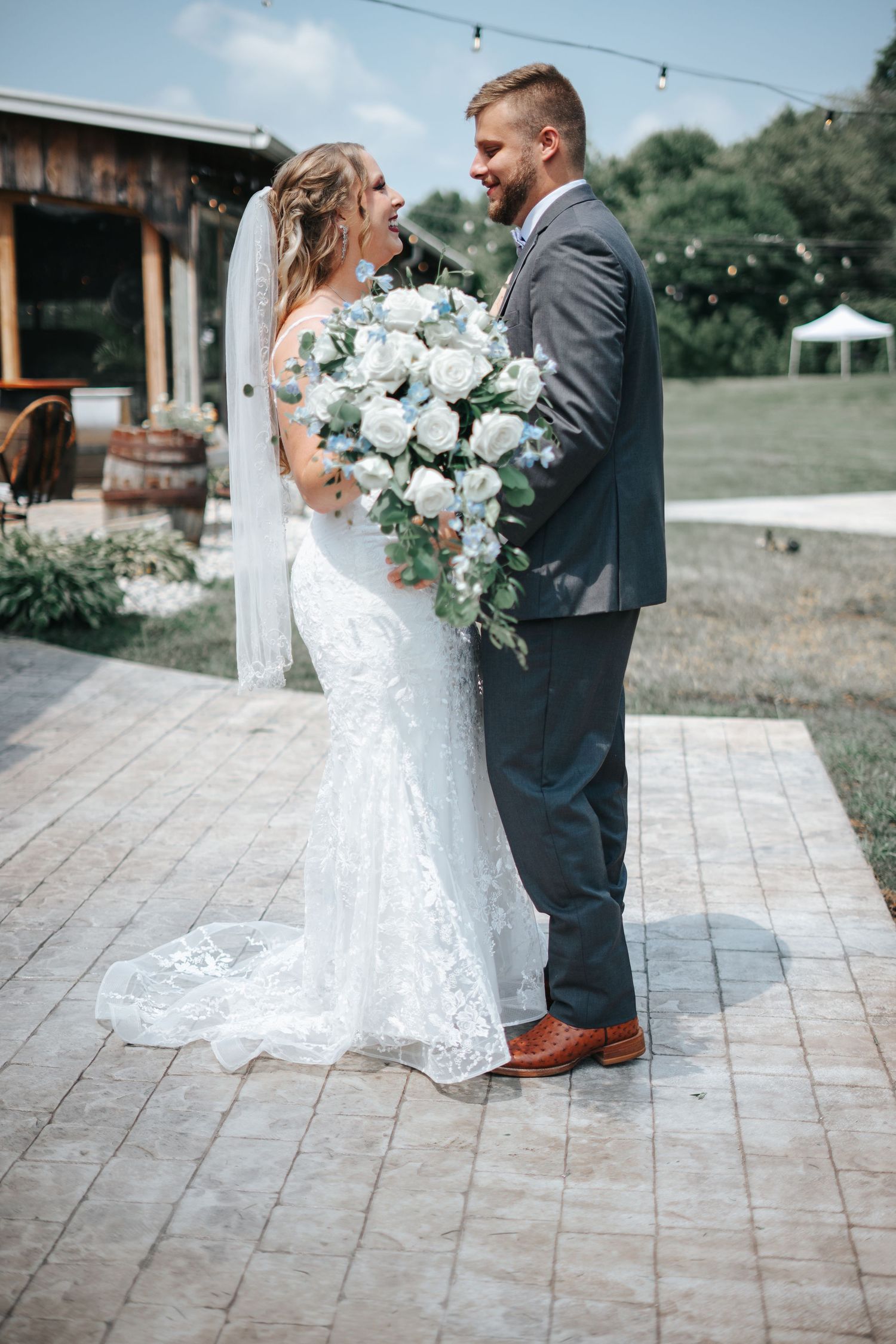 Bride and groom share a joyful moment outdoors, she in lace gown with white bouquet, he in gray suit.