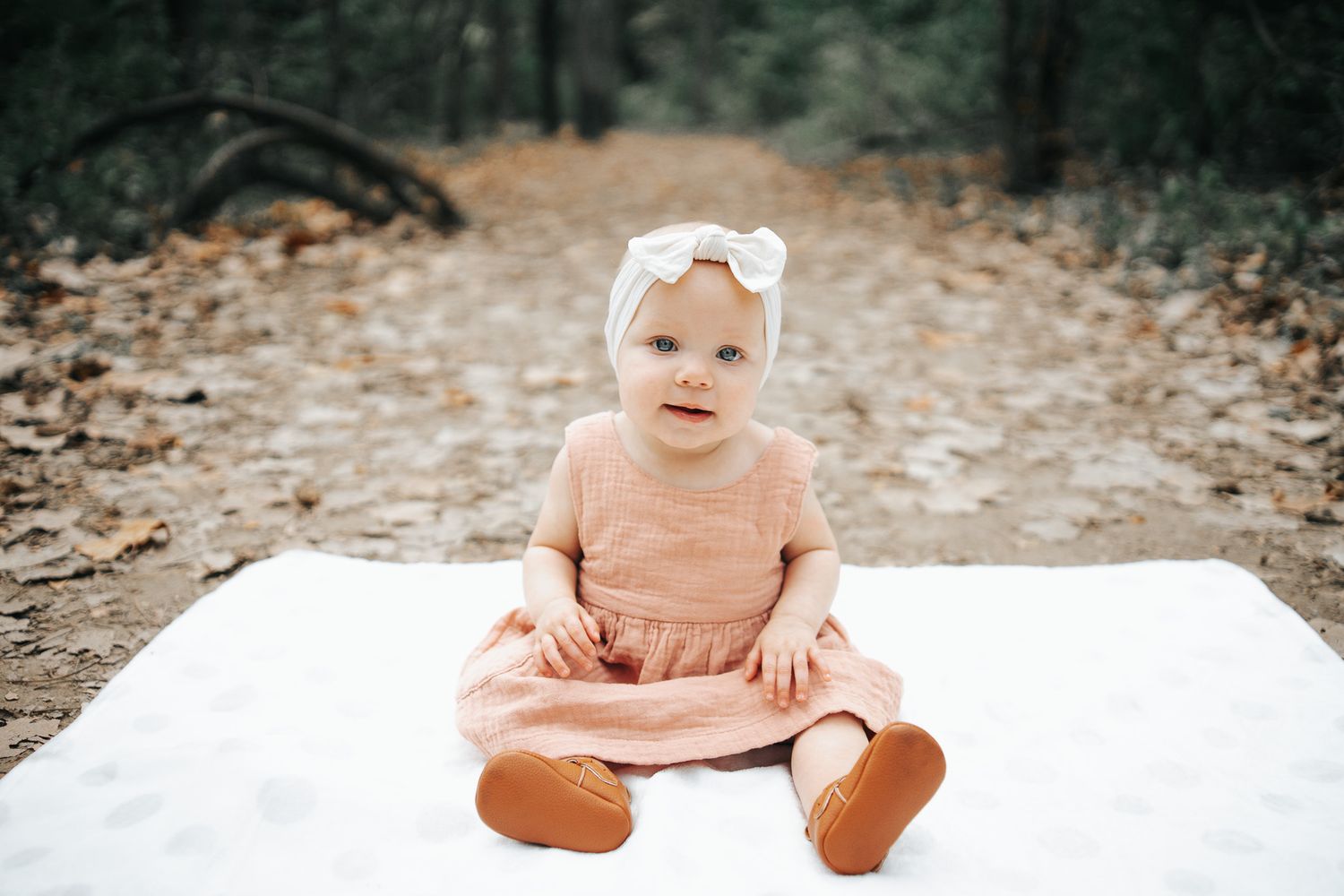 Baby in peach colored outfit sits on white blanket in outdoor setting with natural bokeh background.