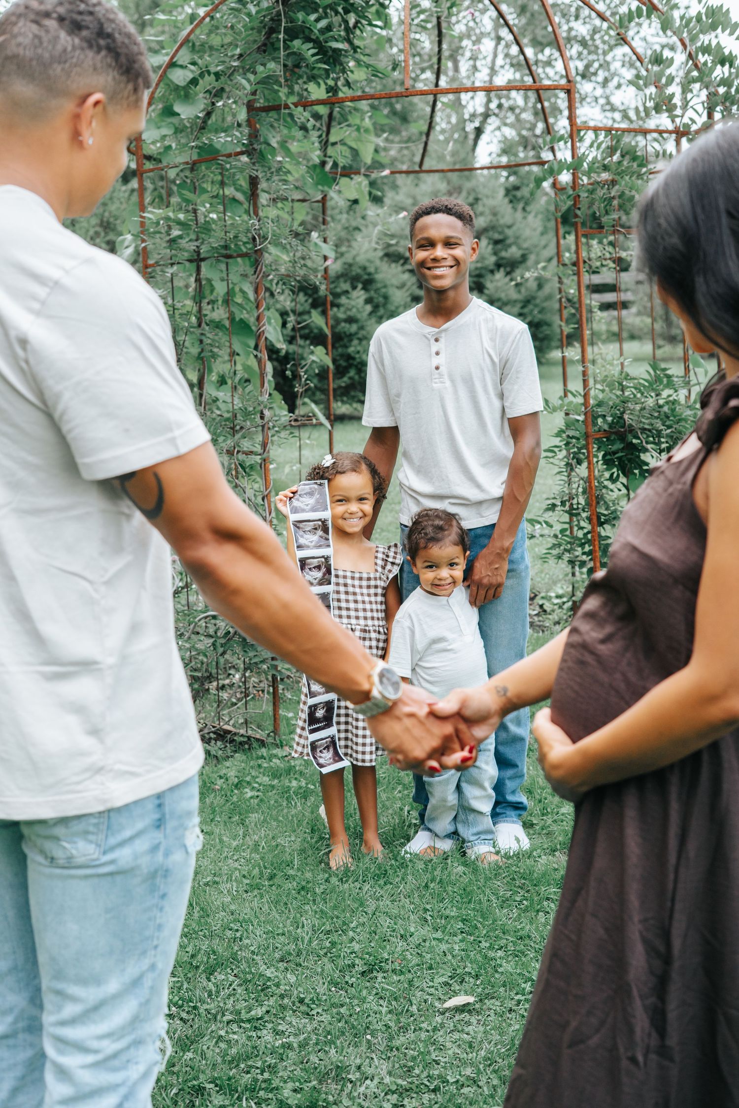 A joyful family holding hands outdoors in a garden, with children smiling near a rustic metal arch.