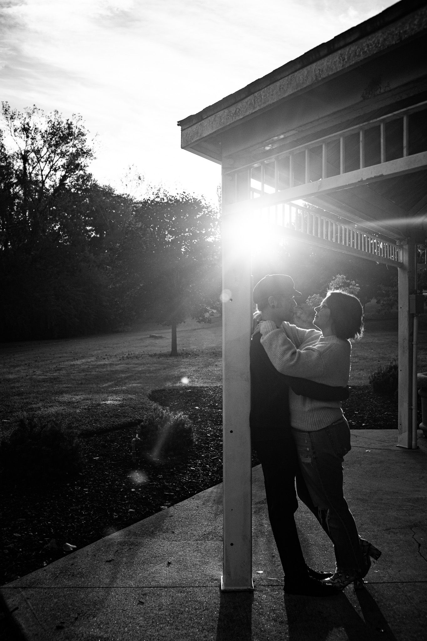 Black and white photo of a couple embracing beside a wooden pavilion post as bright sunlight flares through the trees at sunset.