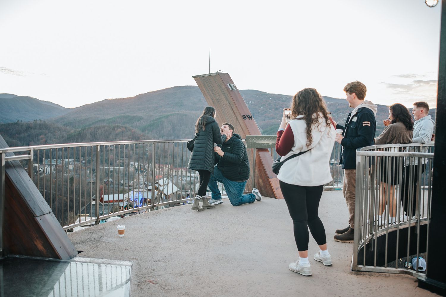 Scenic mountain overlook with visitors enjoying panoramic views from a fenced observation deck on a sunny day.