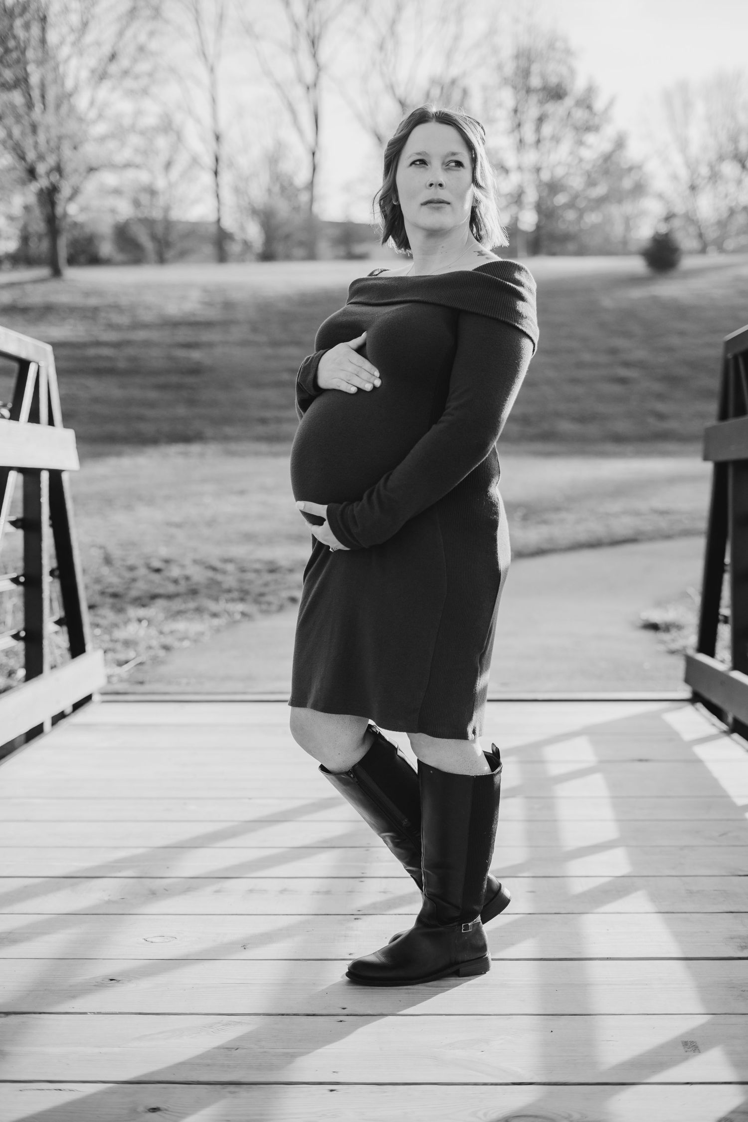 Black and white photo of a pregnant woman in a dress and boots cradling her baby bump on a wooden bridge.