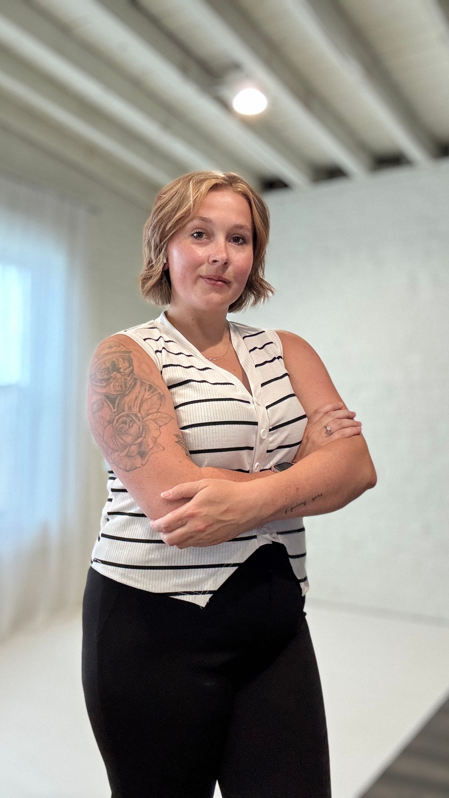 Woman with short wavy blonde hair and a floral arm tattoo stands with crossed arms in a striped sleeveless top in a bright white studio.
