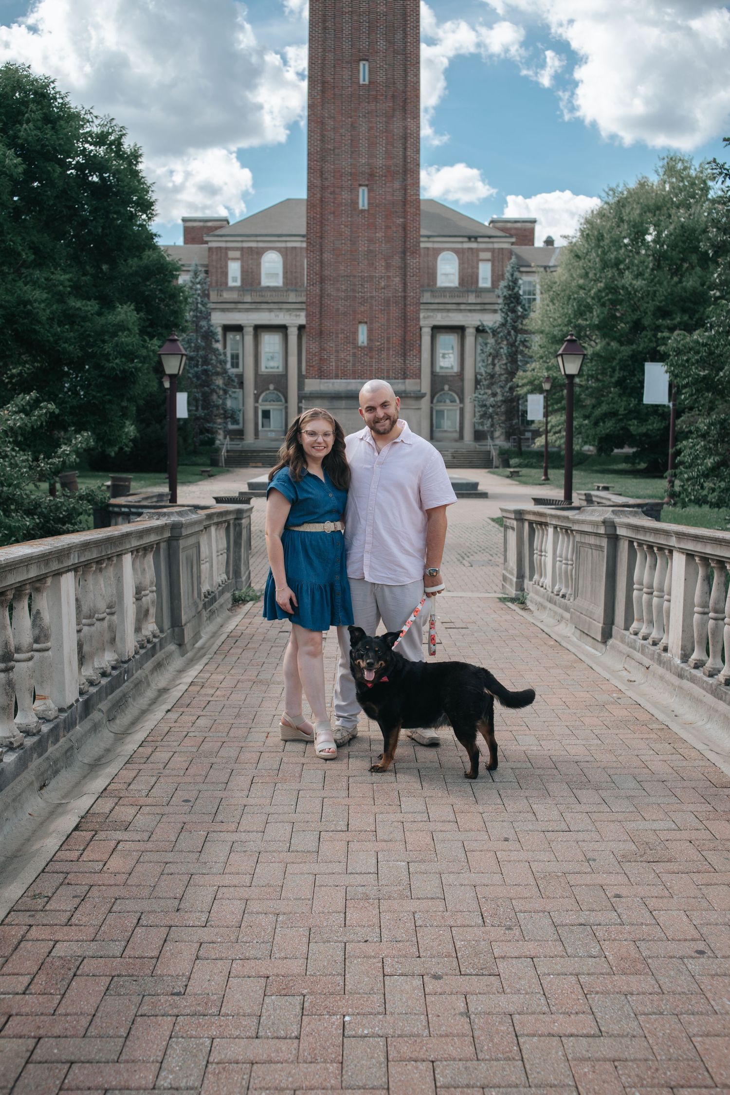 Couple with black dog posing on brick walkway in front of a large brick building on a sunny day.