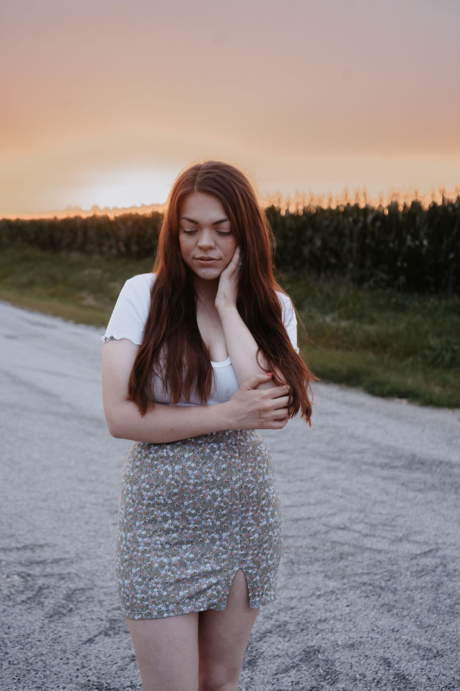 Young woman with long hair in white top and floral skirt poses on rural road at sunset.