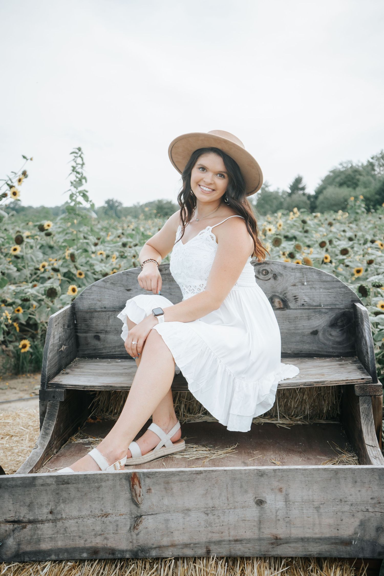 Young woman in white dress and tan hat sits smiling on wooden wagon in sunflower field.