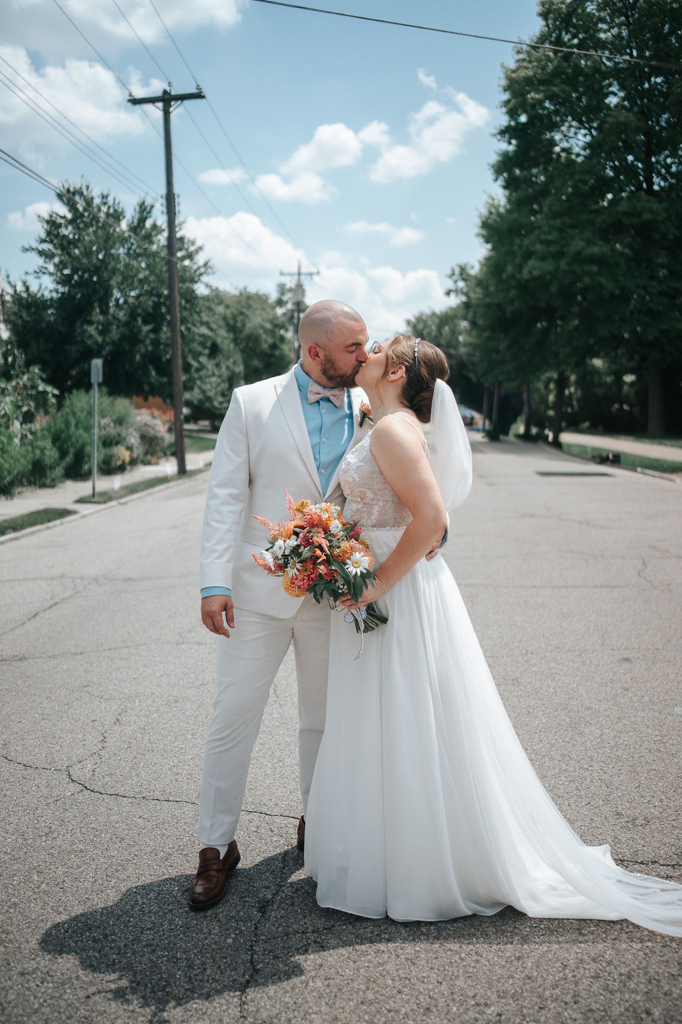 Bride and groom sharing a kiss on a sunny street, bride holding colorful bouquet in white gown, groom in white suit.