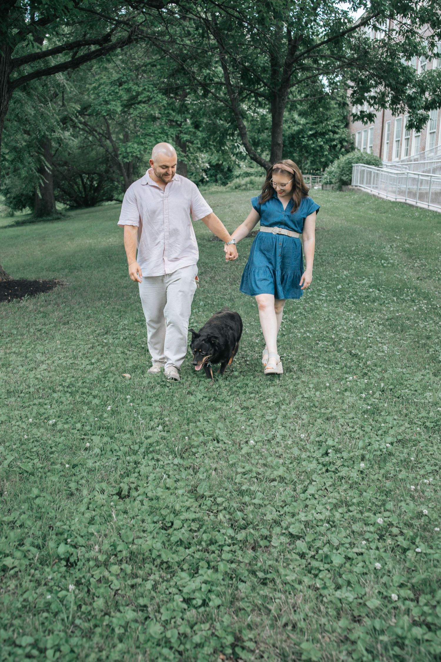 Couple holding hands walking black dog on green grass park, man in pink shirt, woman in blue dress.