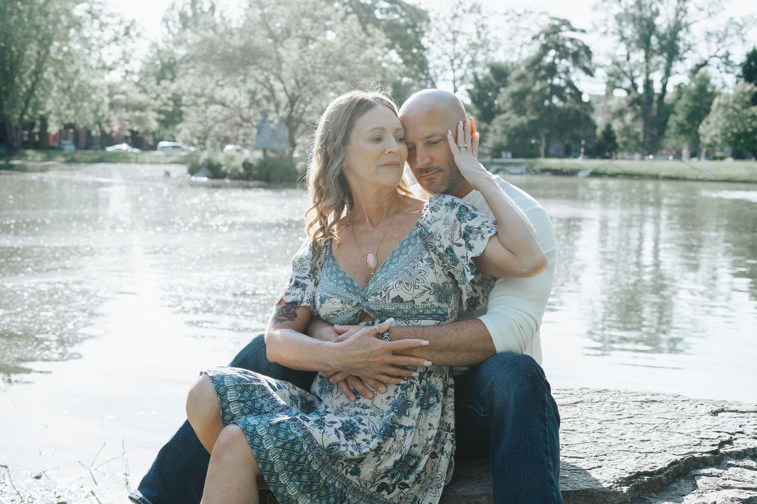 A couple embraces lovingly by a serene lake surrounded by trees in a romantic outdoor spring setting.