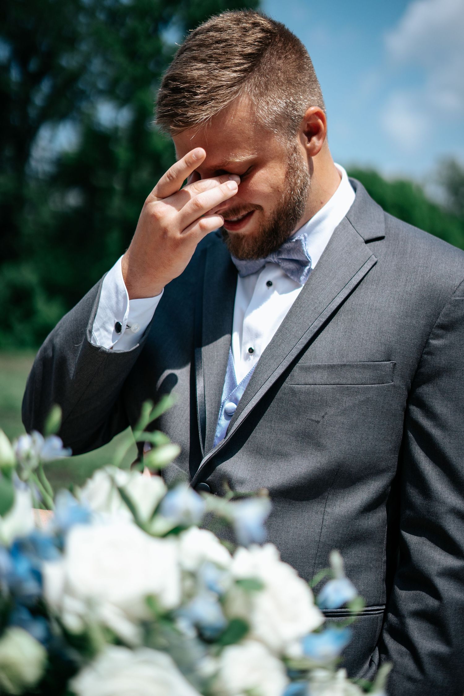 Emotional groom in a gray suit and bow tie wipes his eyes with his hand beside a bouquet of white and blue flowers outdoors.