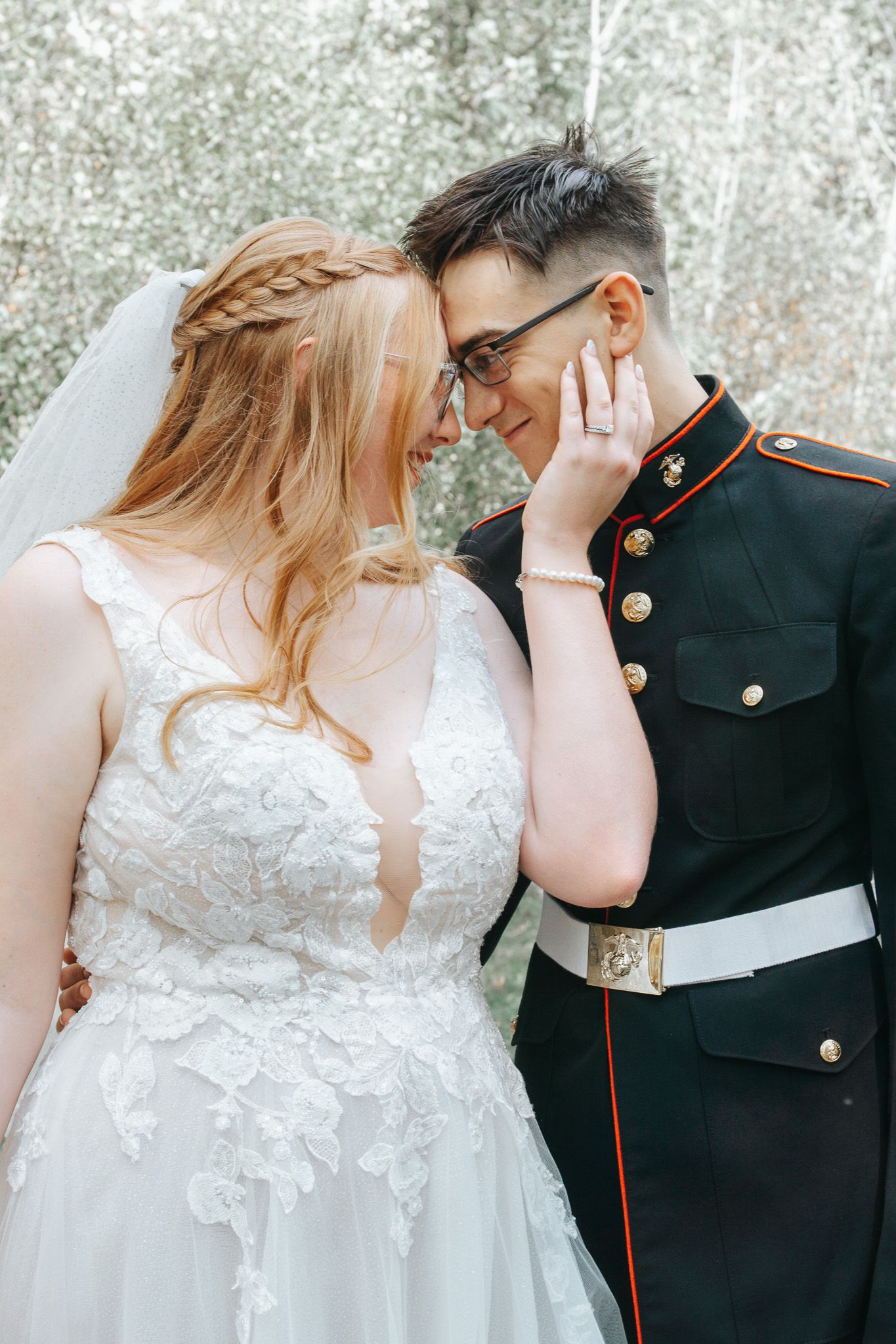 A bride in a white lace wedding gown with a braided hairstyle touches foreheads with her groom, who wears a dark military dress uniform.
