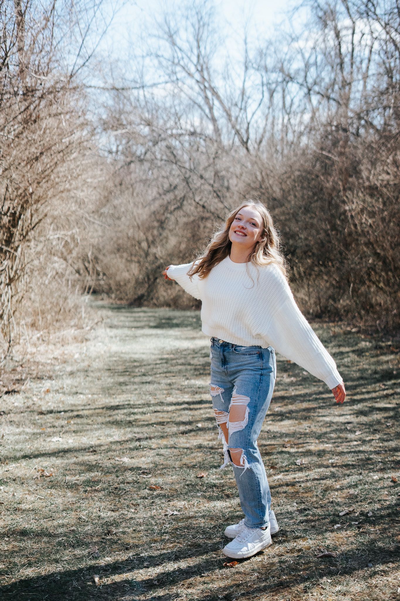 A person in a white sweater and ripped jeans dances playfully outdoors on a sunny winter day.