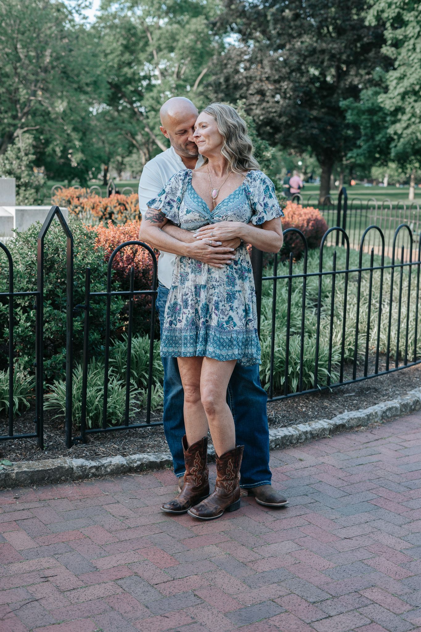 A couple embraces on a brick pathway near a decorative iron fence and garden with the woman wearing a blue dress and cowboy boots.