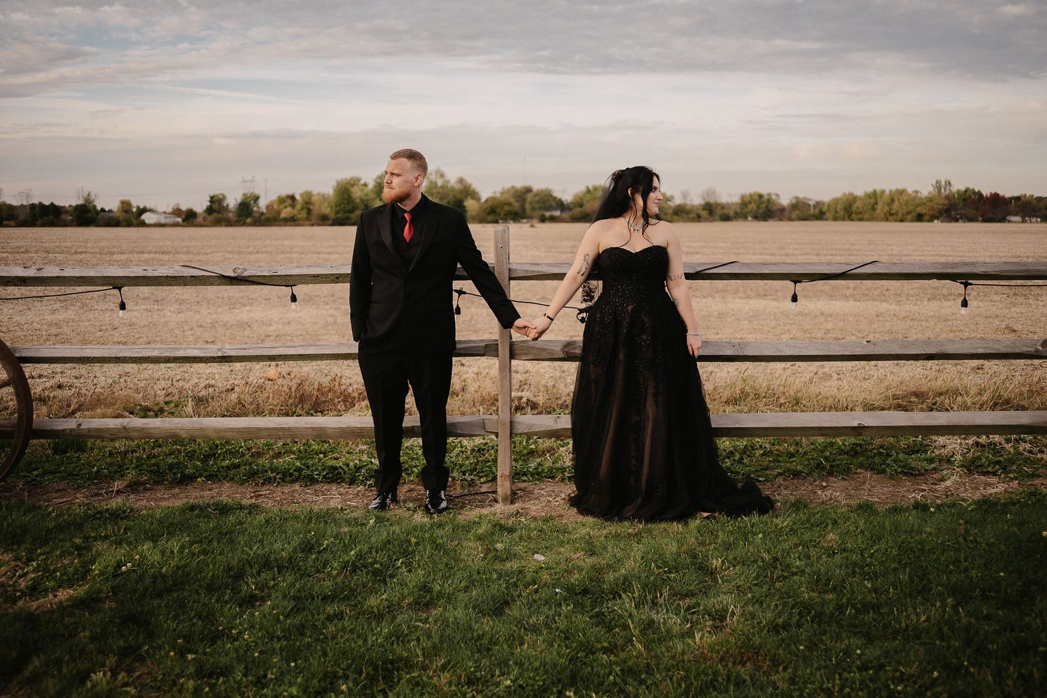 A couple stands together in a grassy field at sunset with a dramatic cloudy sky in the background.