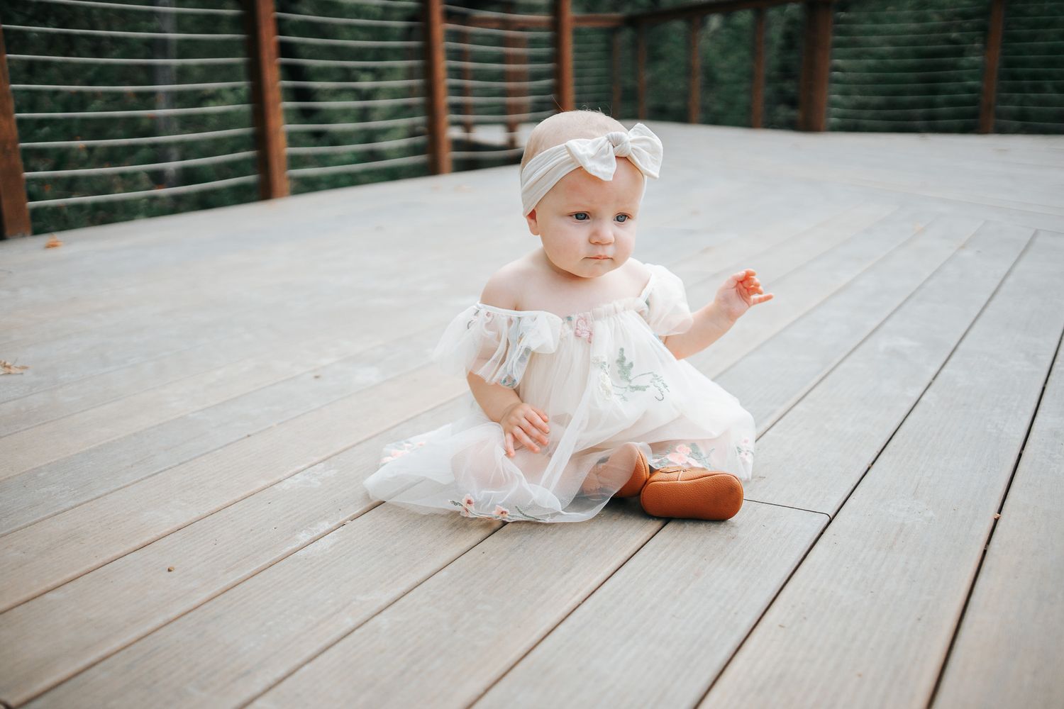 A baby in a white lace dress sits and crawls on a wooden deck in a series of connected photos.