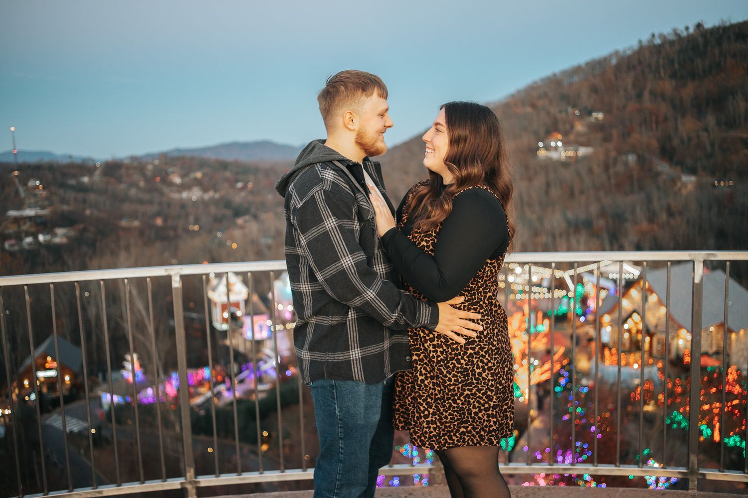 A couple shares a romantic moment on an observation deck overlooking a twinkling city at dusk.