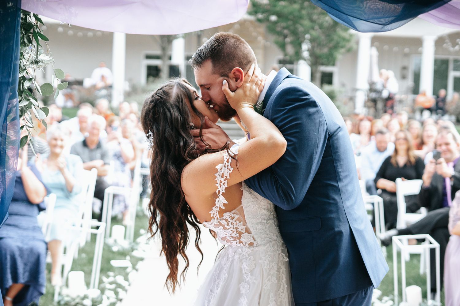 Bride in a lace wedding gown and groom in a navy suit share a romantic kiss while smiling guests watch at an outdoor ceremony.