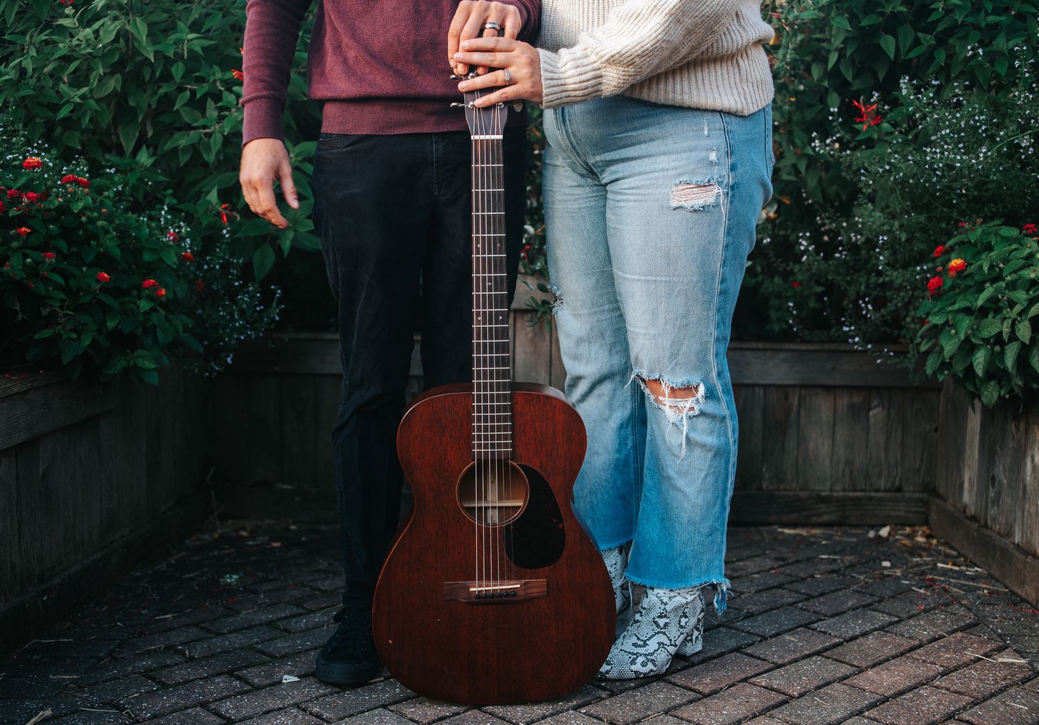 Couple standing close together behind a mahogany acoustic guitar on a brick patio, surrounded by green shrubs with red flowers.