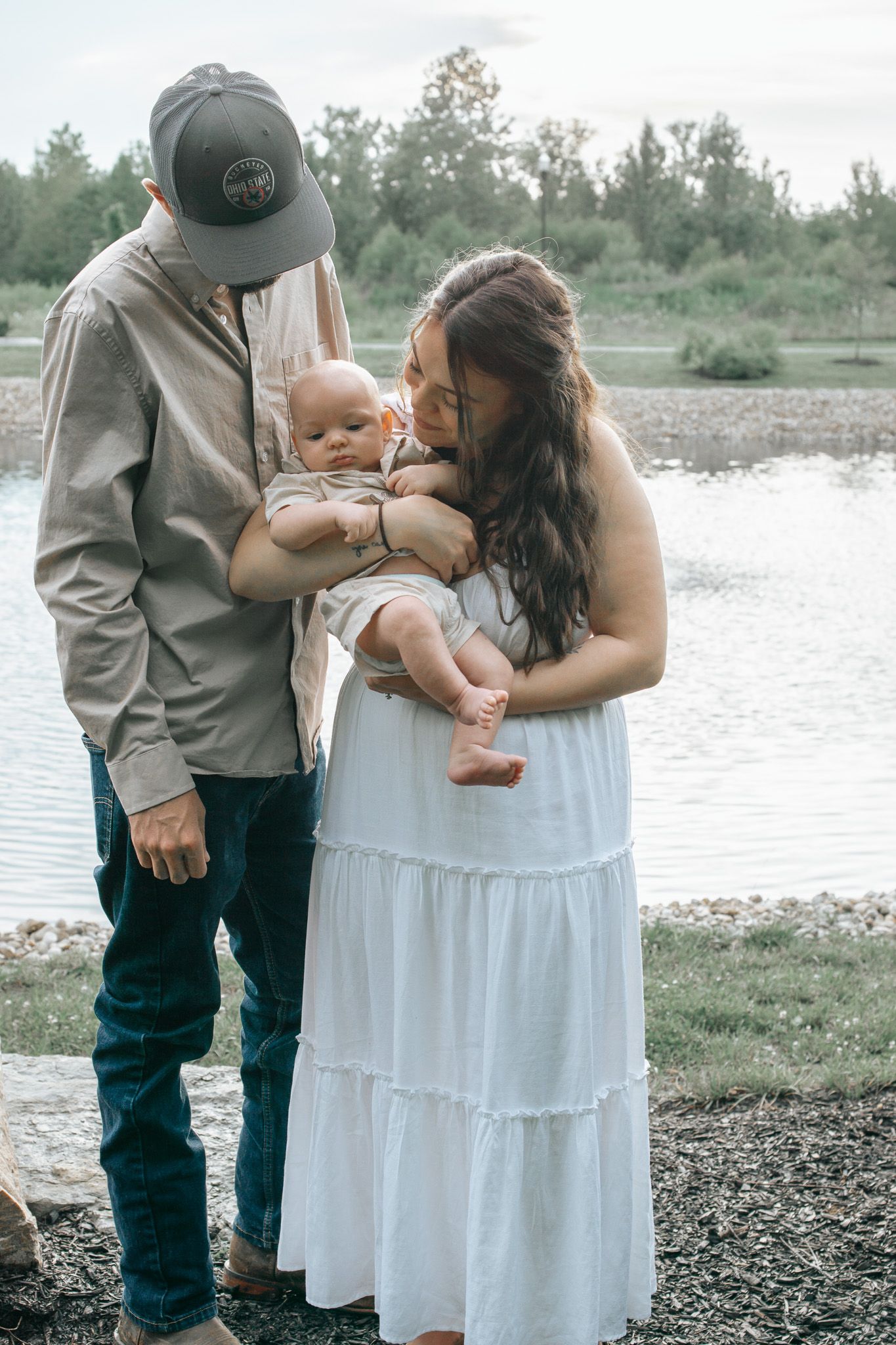 A young couple in casual attire lovingly gazes at their newborn baby by a serene lakeside setting.