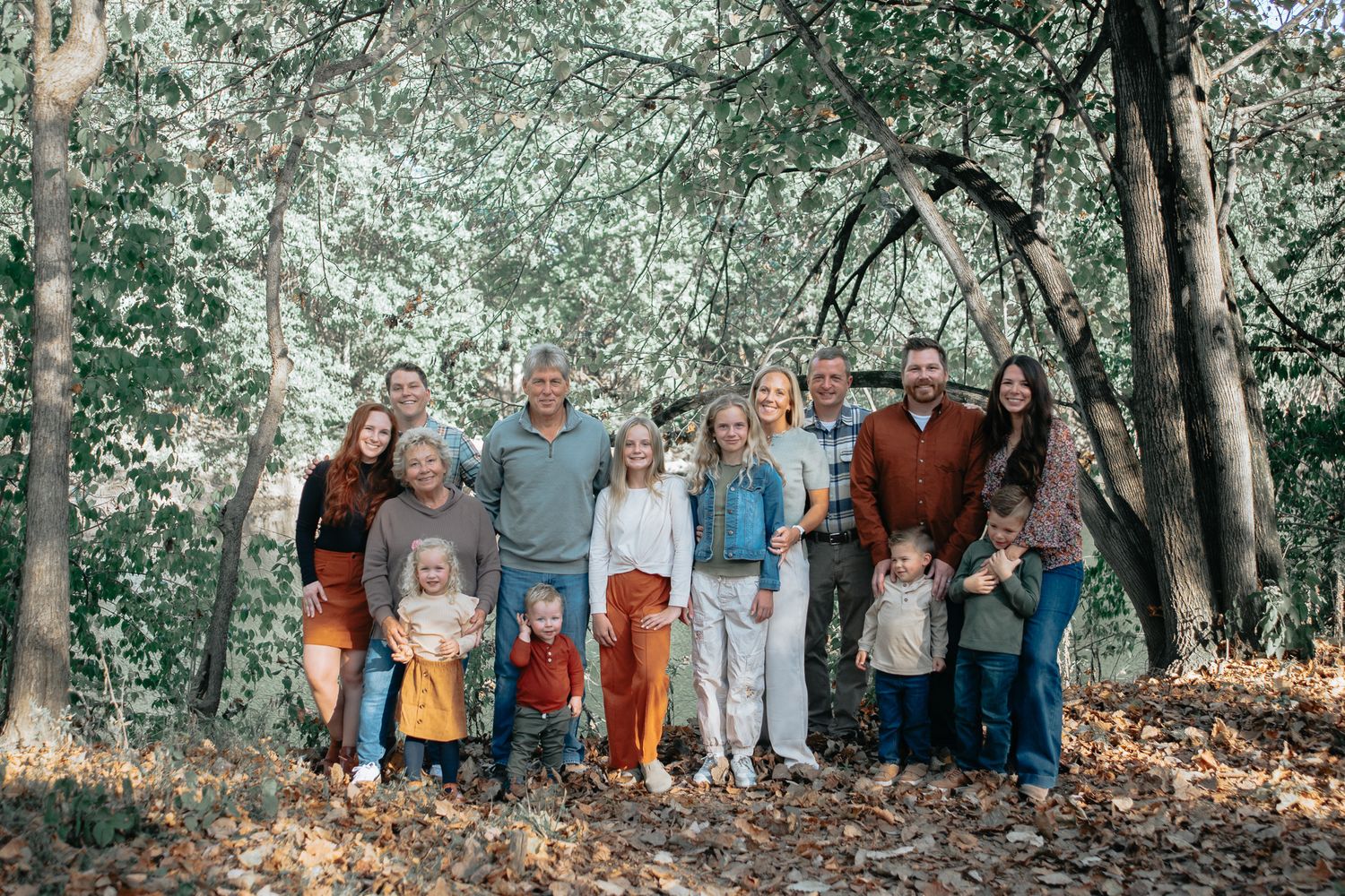 A large group of people stand together in a wooded outdoor setting during a photo session.