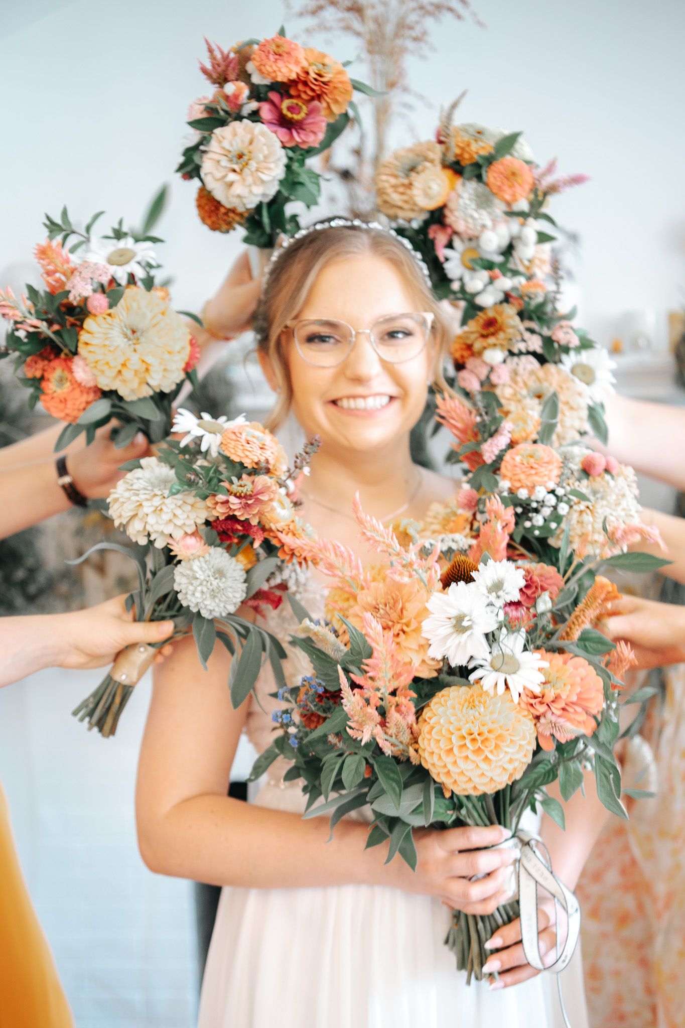 Smiling bride in glasses holds orange floral bouquet, surrounded by bridesmaids bouquets and flower arch backdrop.