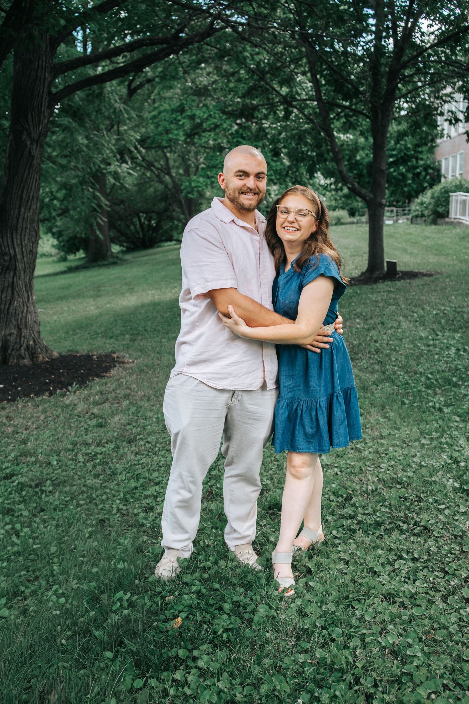 Smiling couple embracing outdoors in a lush green park, man in pink shirt and woman in blue dress.