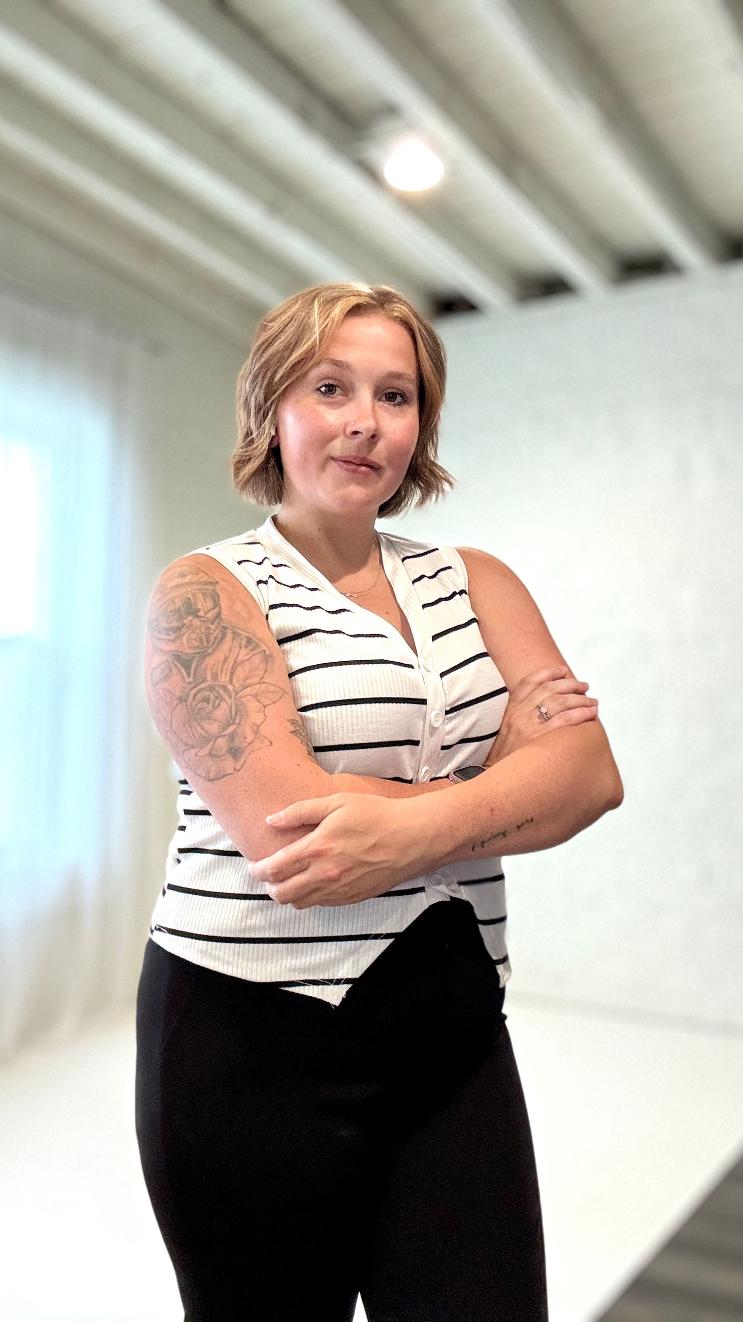 Person in a striped white top and black pants poses against a white wall showing a visible arm tattoo.