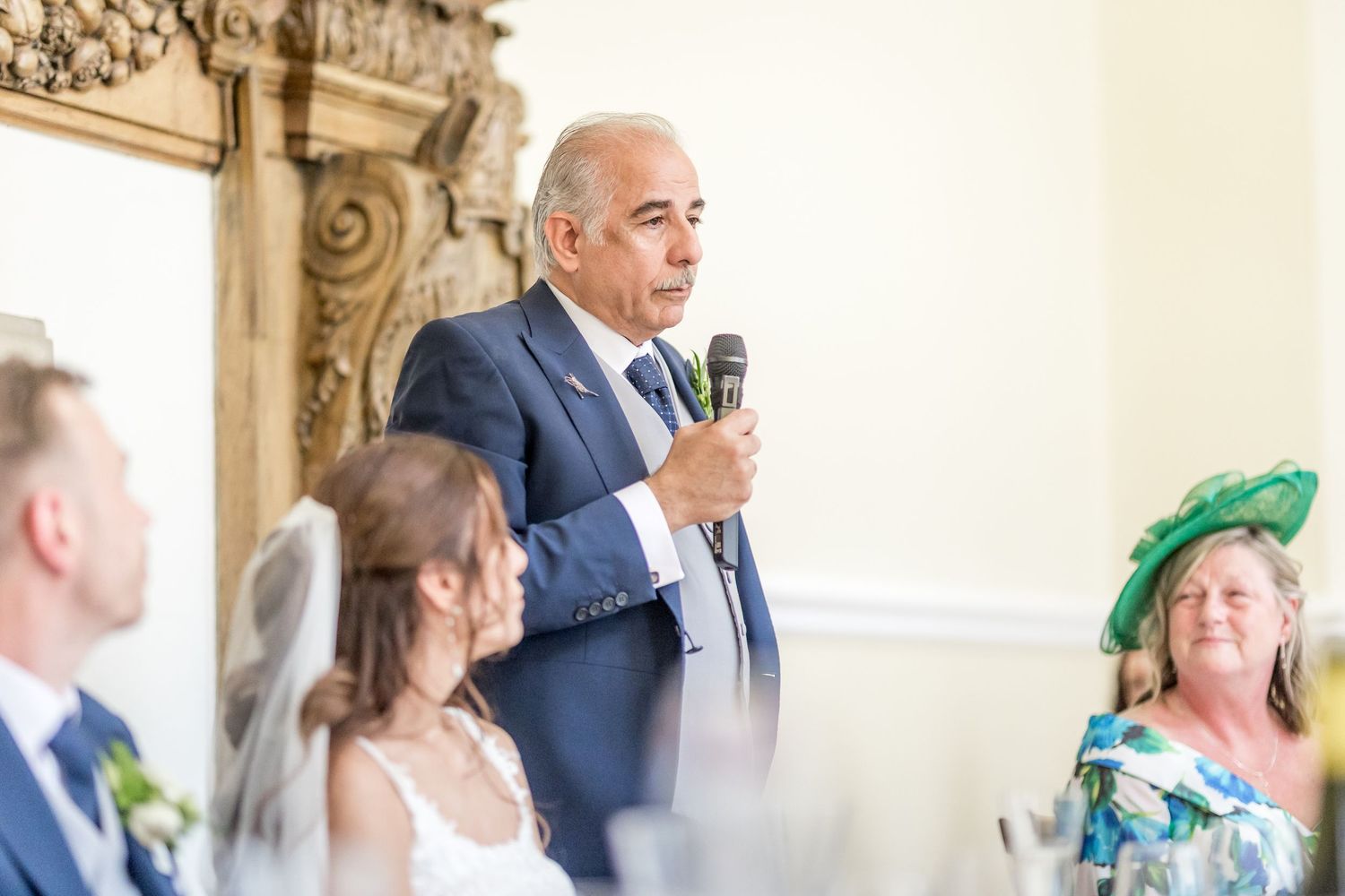 Wedding reception guests listen attentively to speeches in an elegant venue with ornate decor.
