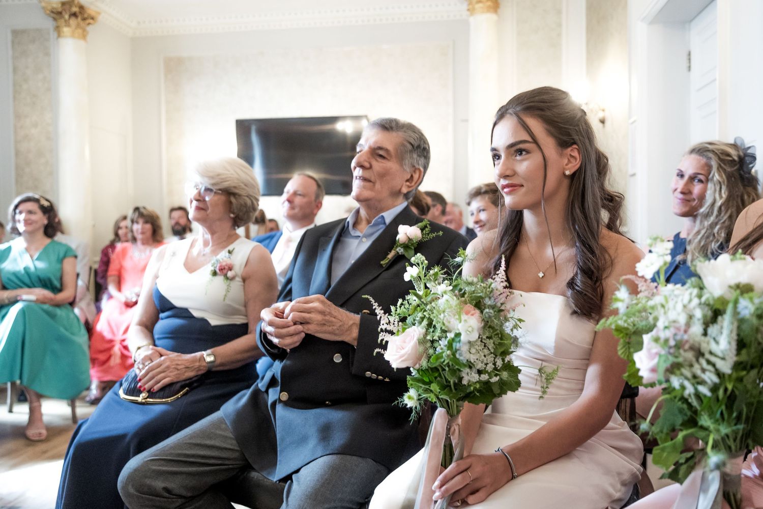 Wedding guests and couple seated in bright ceremony room with white floral decorations.