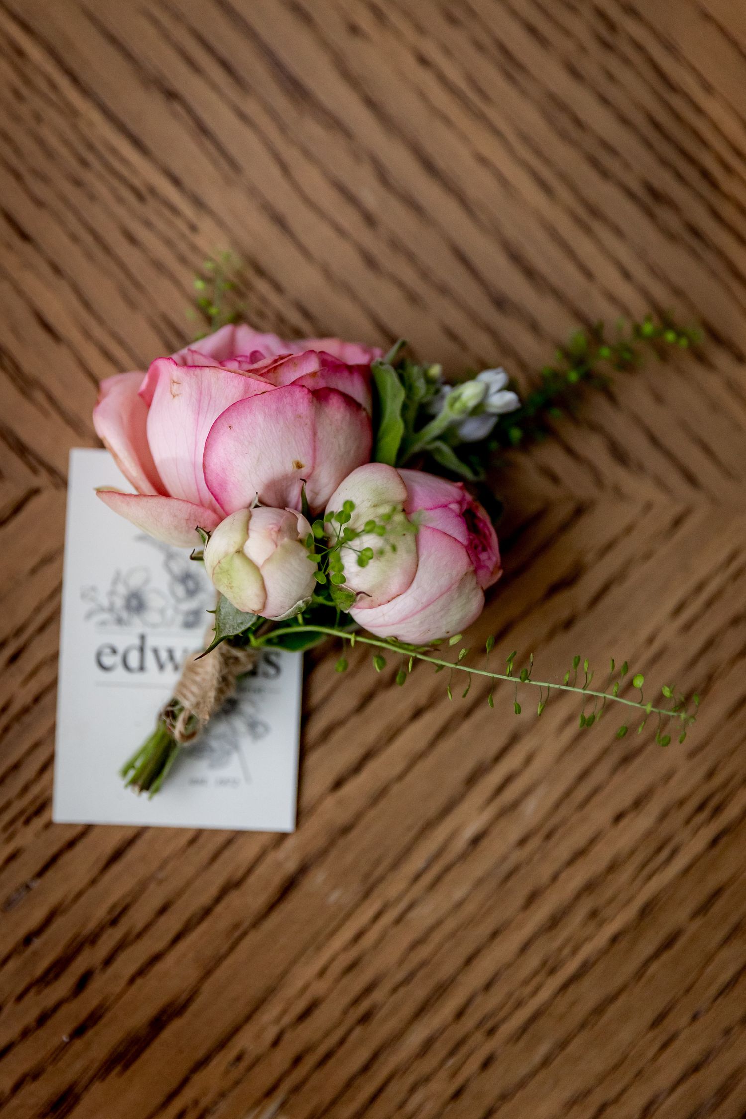Pink roses boutonniere with greenery lying on wooden surface next to a card.