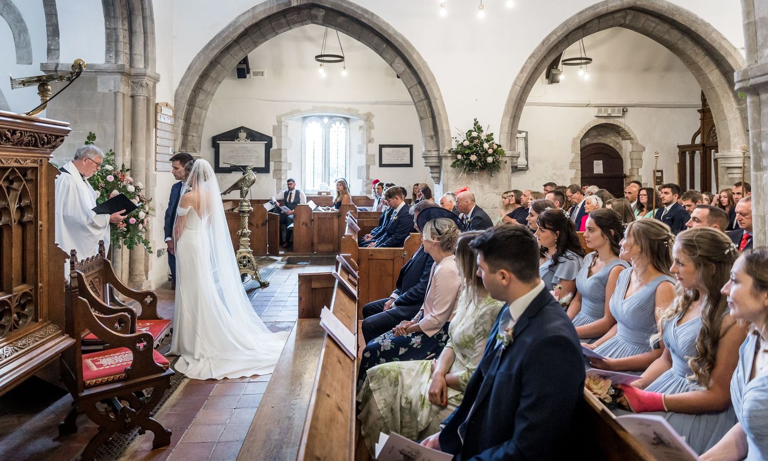Wide angle view of church interior with guests seated in wooden pews during wedding ceremony.