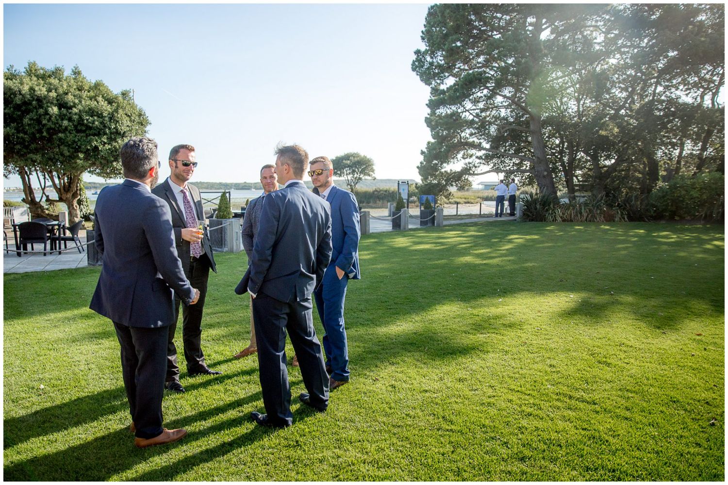 Wedding party gathered on a lush green lawn during golden hour with sunlight streaming through trees in the background.