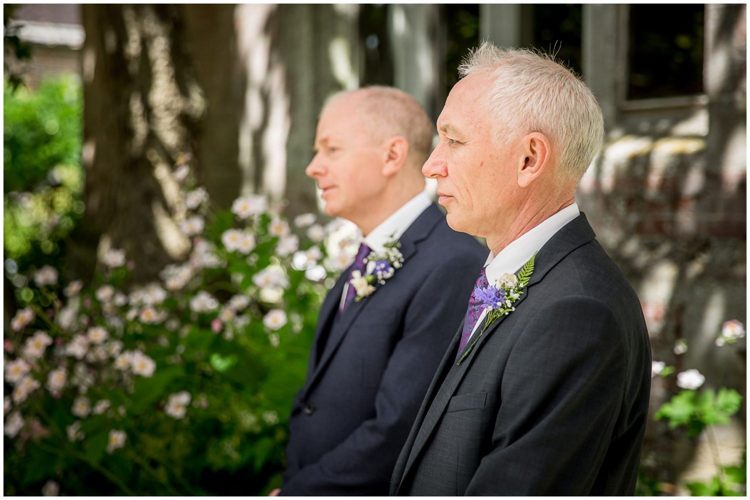 Two men in formal attire stand close together during an outdoor ceremony with trees in the background.