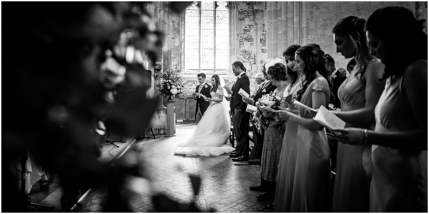 A black and white panoramic wedding ceremony photo showing guests standing in a church with sunlight streaming through windows.