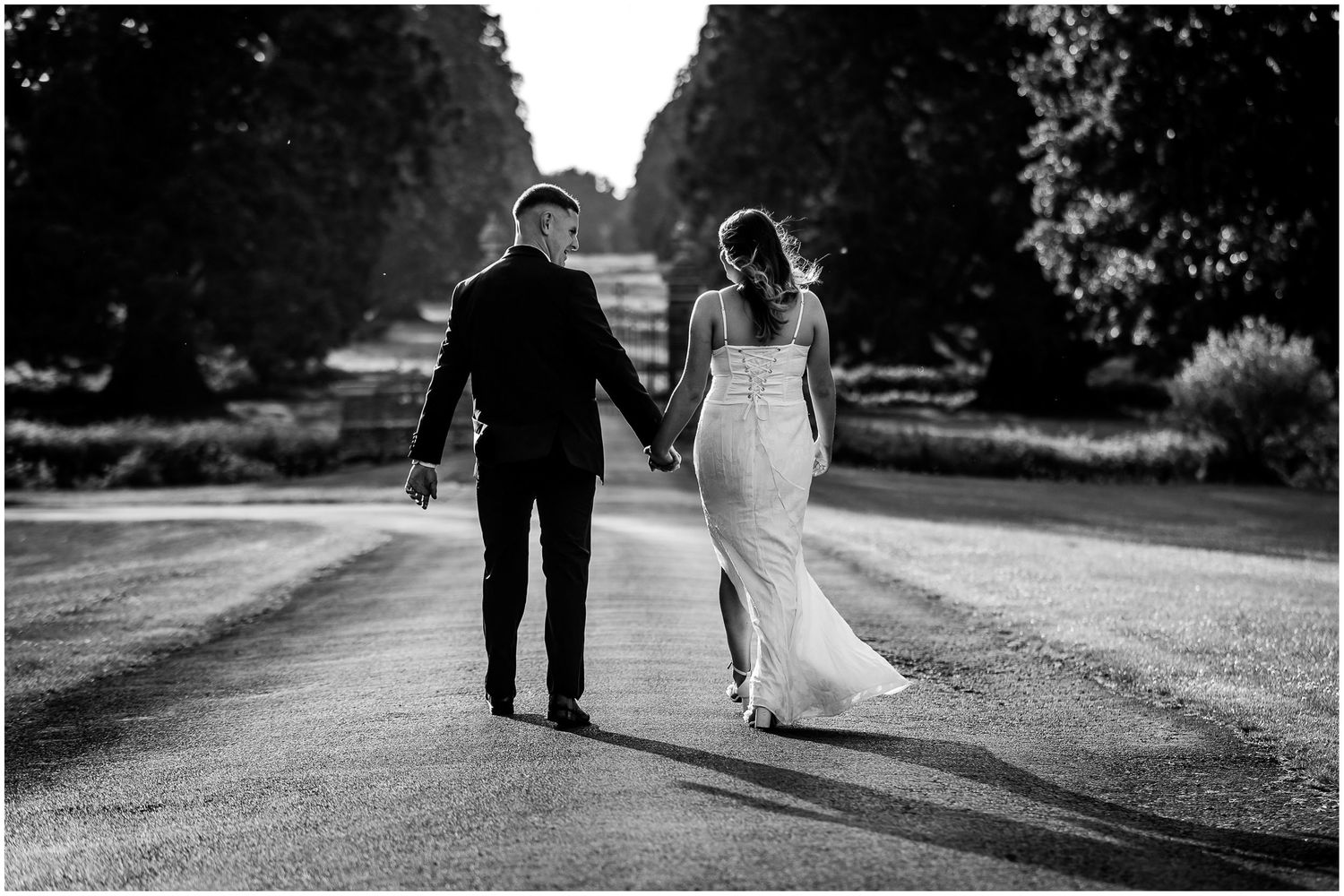A couple in wedding attire walks down a tree-lined path holding hands in black and white photography.