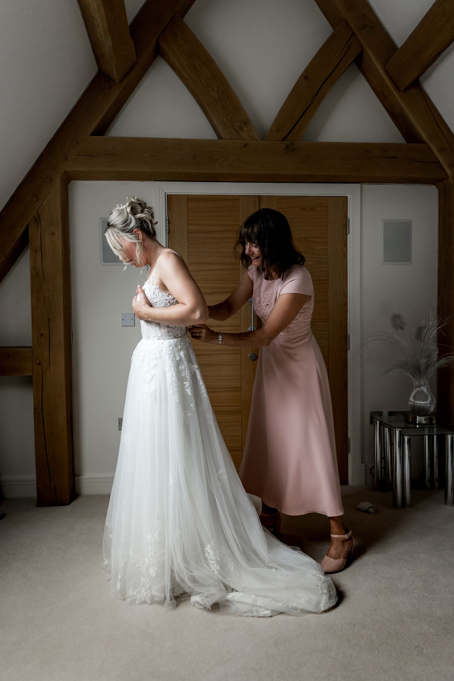 A person in a pink dress helps adjust the hem of a white wedding gown in a room with exposed wooden beams.