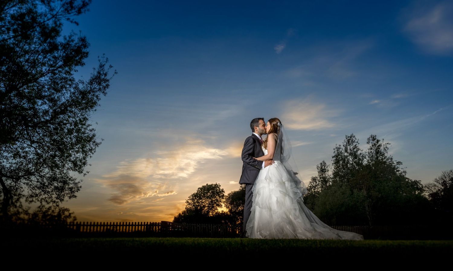 A romantic silhouette of a couple in wedding attire embracing against a dramatic sunset sky with trees in the background.