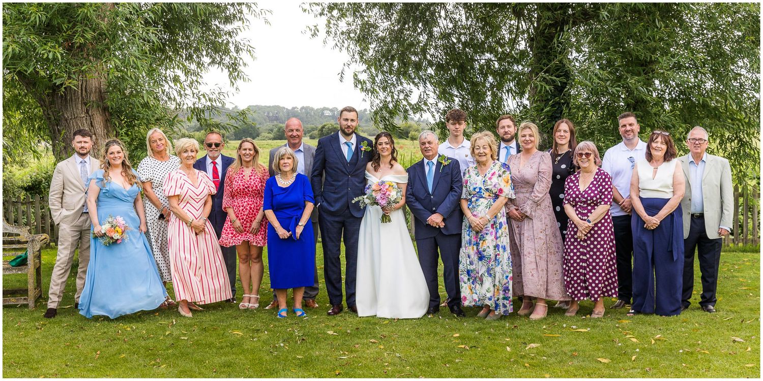 Larger group wedding photograph in the shade of the willow trees lining the river in the gardens of Sopley Mill in the New Forest