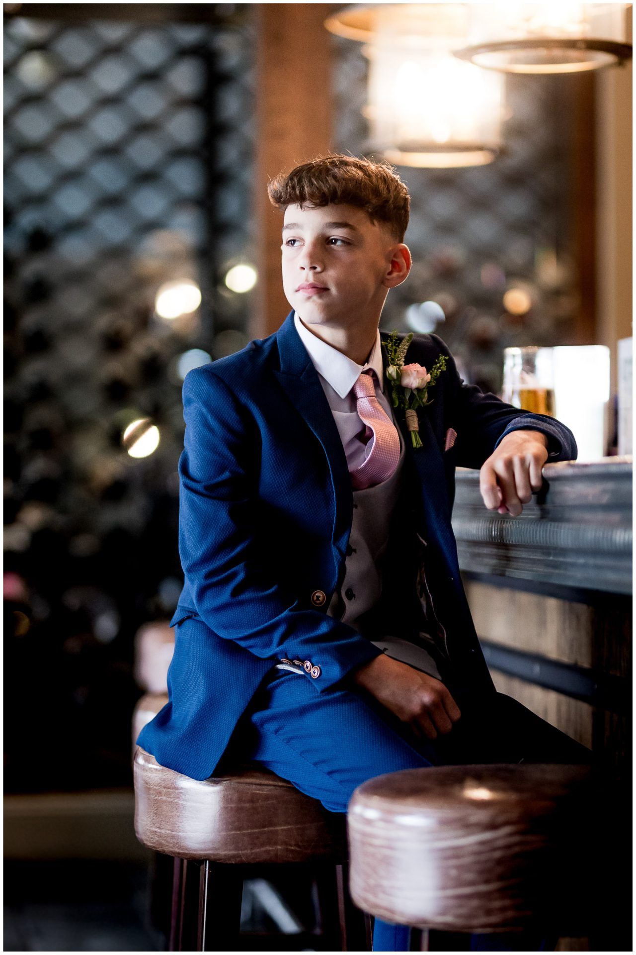 Young person in blue suit with pink tie sits at bar counter with boutonniere.
