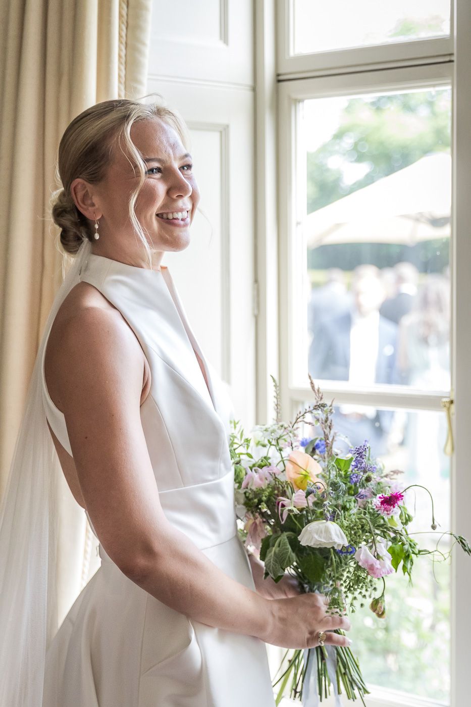 Bride in elegant white dress holding colorful wildflower bouquet by sunlit windows.