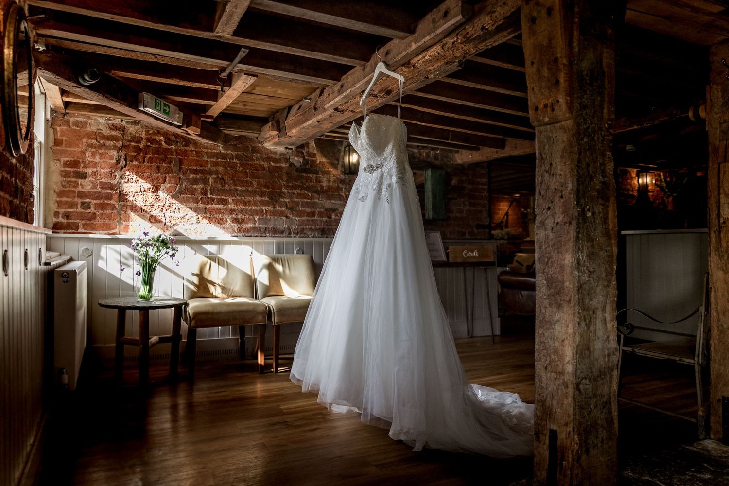 White wedding dresses hang from wooden beams in a rustic brick venue interior.