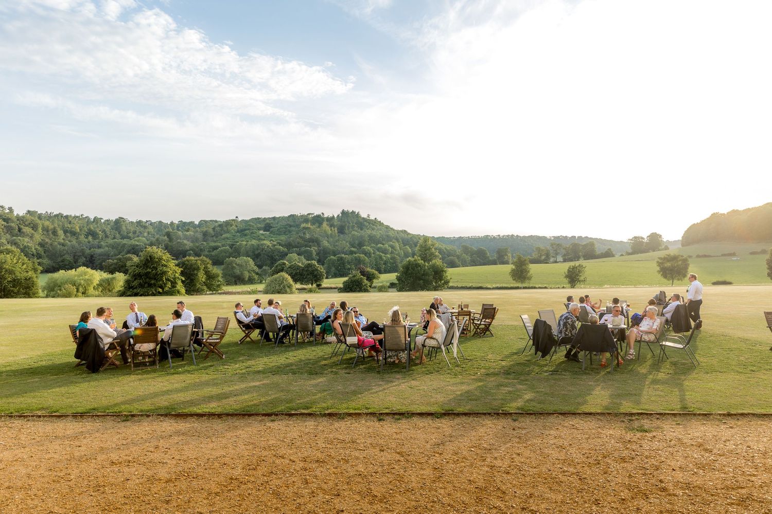 Panoramic view of wedding guests seated at tables on a grassy hillside under blue skies.