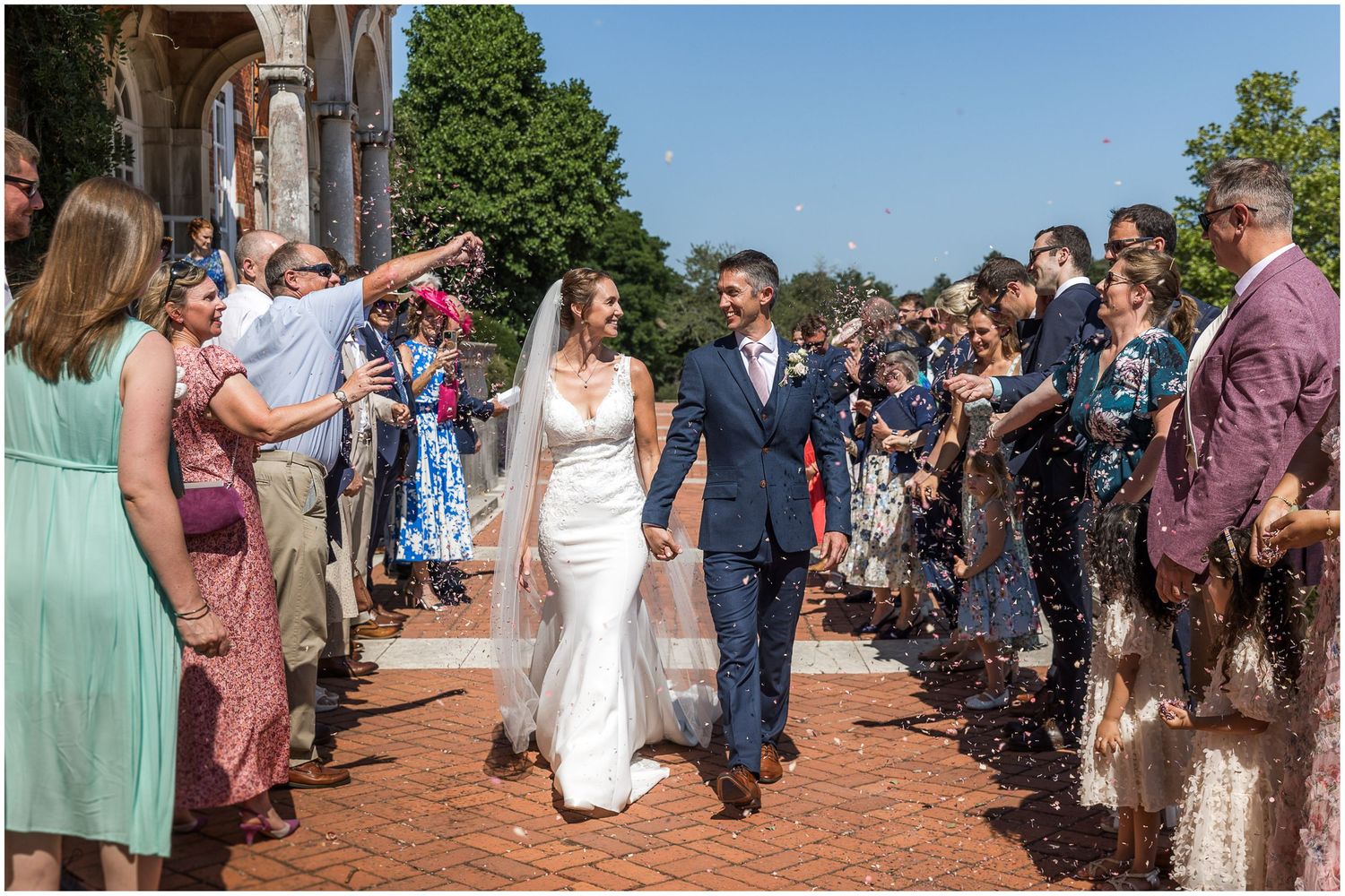 Bride and groom walk through confetti shower surrounded by smiling guests outside Walhampton School on their wedding day.