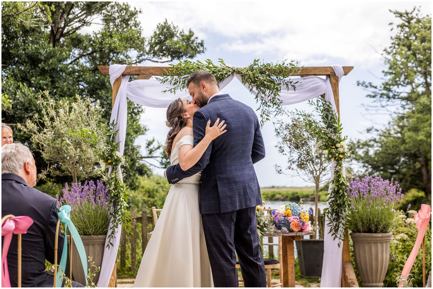 Bride and groom share first kiss under floral wooden arch at Sopley Mill, in the riverside outdoor ceremony space